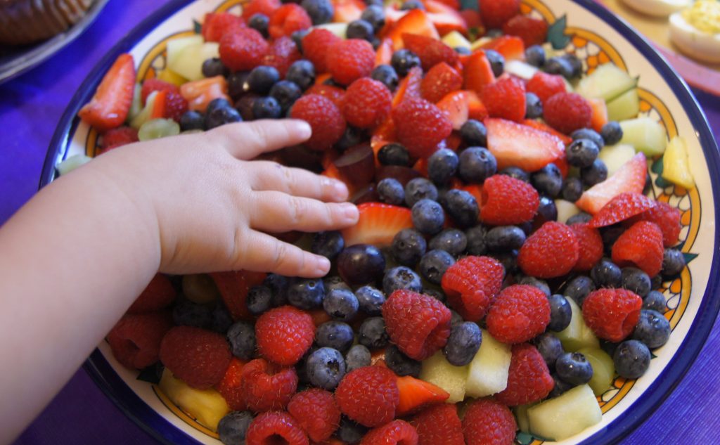 Una variedad de frutillas en un bol grande, y la mano de un niño agarrando las frutas.