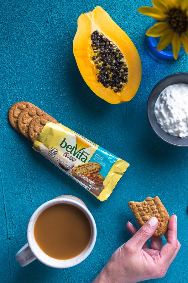 biscuit belvita en la mano ya mordido, el paquete abierto con tres biscuits, papaya, queso requesón, una taza de café.
